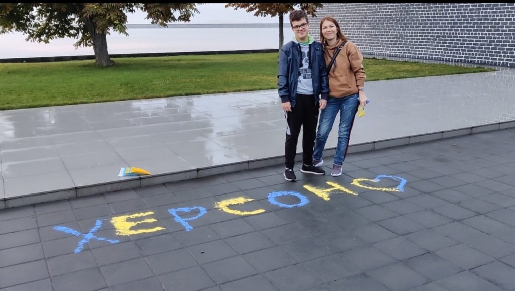 Two persons standing over Kherson written in blue and yellow on the road, in Cyrilic