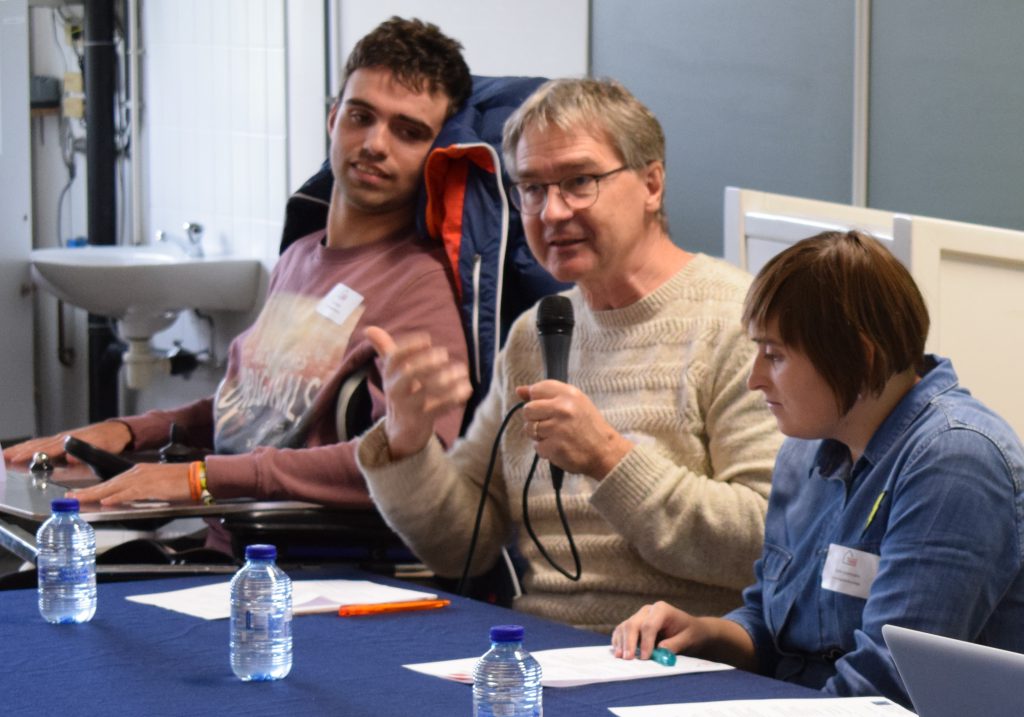 Three people sitting at a table. The one in the middle holds a microphone.