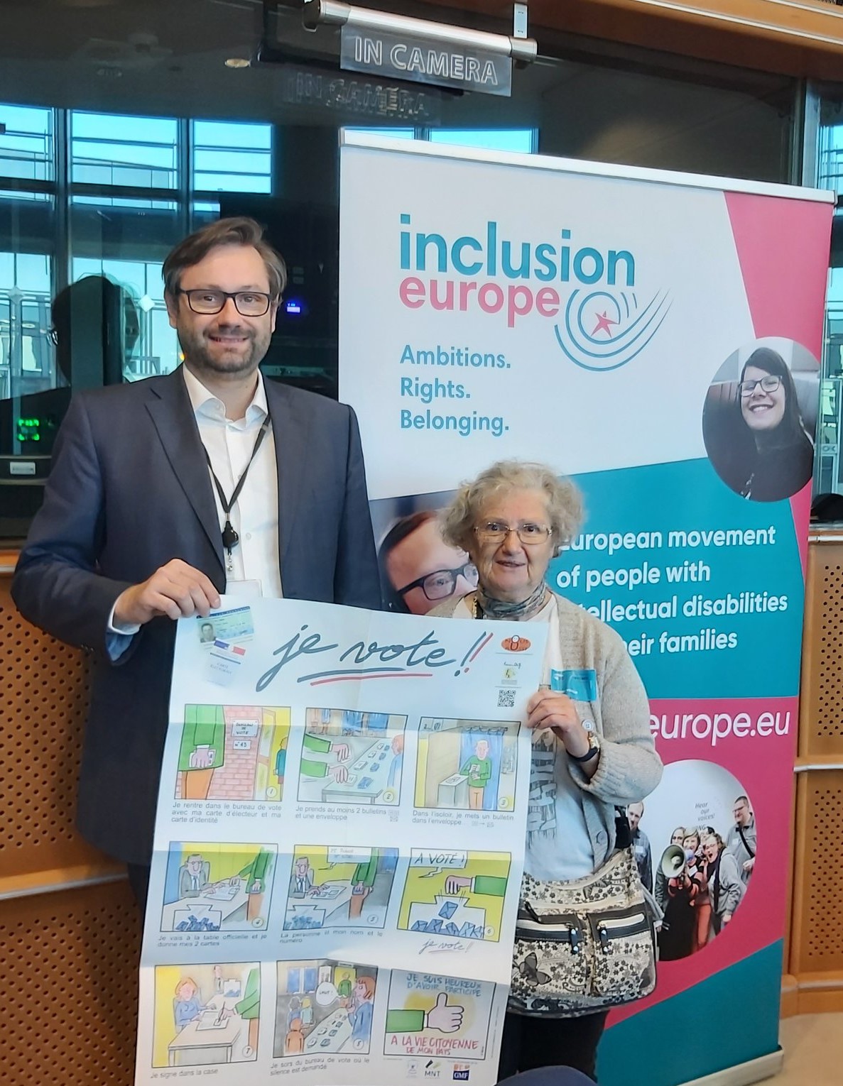 A tall man with glasses and an older woman with glasses standing in front of a banner, holding a poster